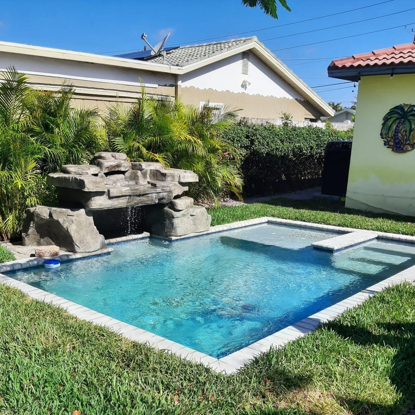 Residential backyard swimming pool with rock waterfall feature, shallow sun shelf, and tropical landscaping.