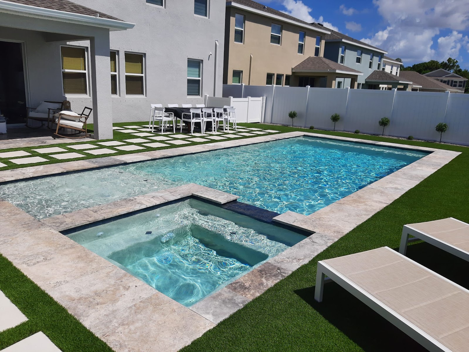 Modern backyard swimming pool with integrated spa, stone coping, and artificial turf landscaping behind a residential home.