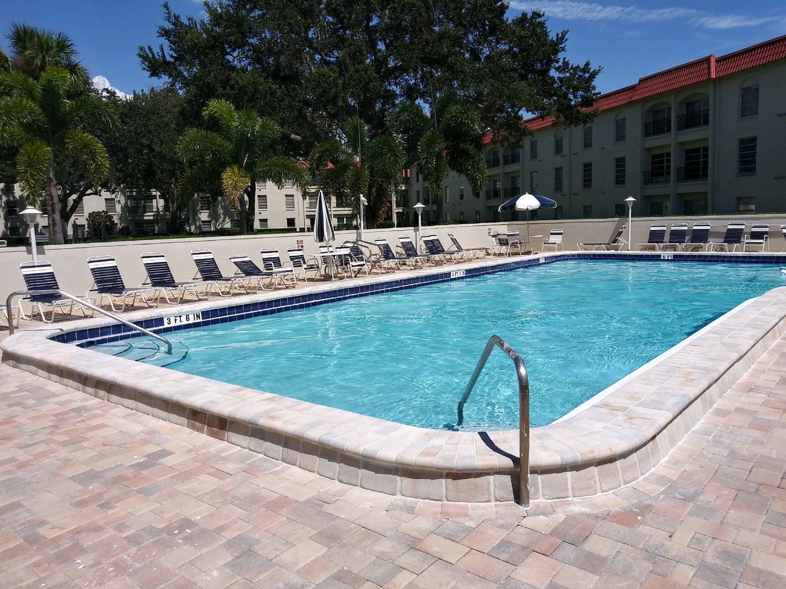 Outdoor community swimming pool with clear blue water, lounge chairs, and surrounding apartment buildings under a sunny sky.