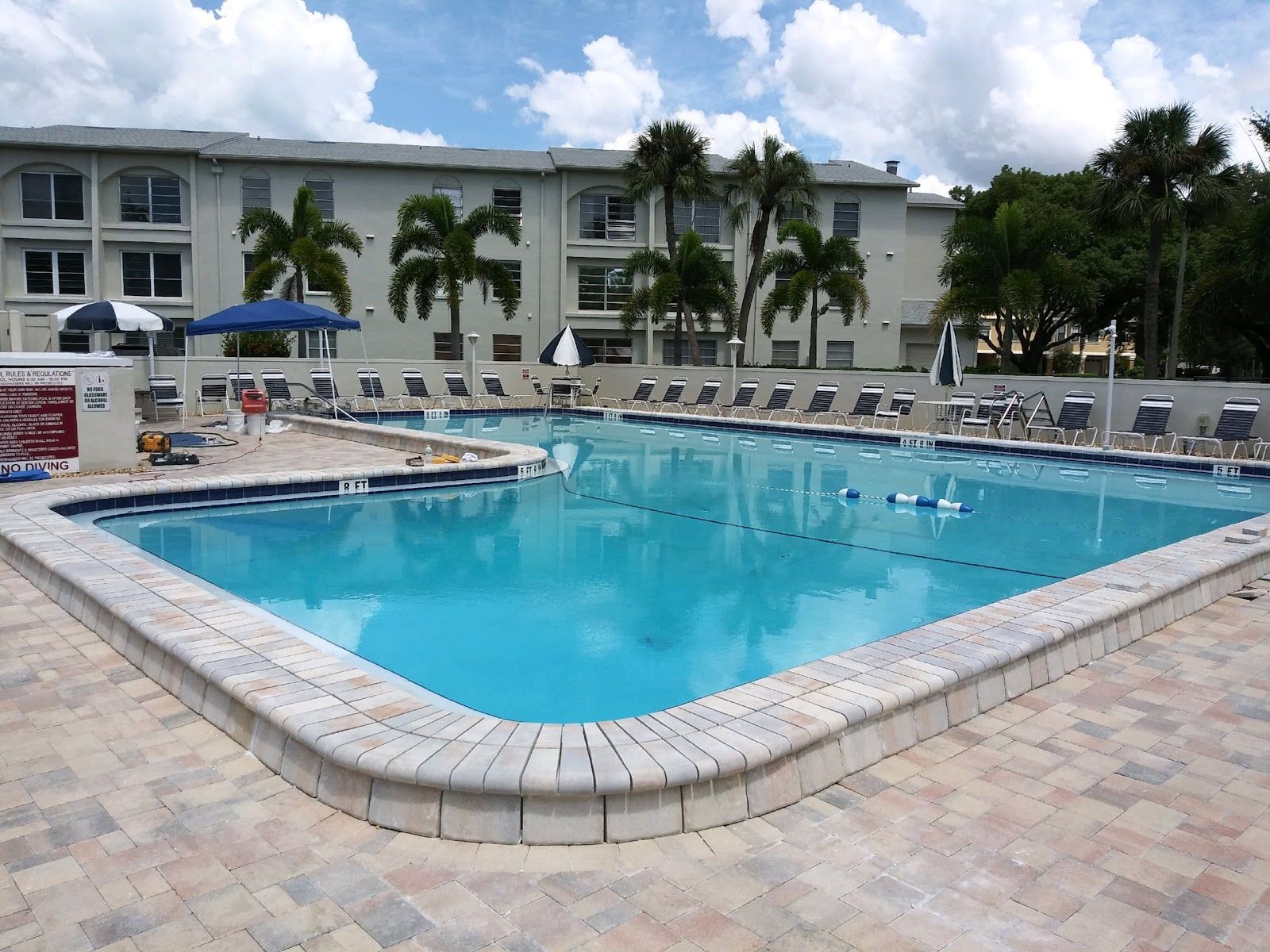Large community swimming pool with light stone paver deck, lounge chairs, and palm trees in a residential complex.