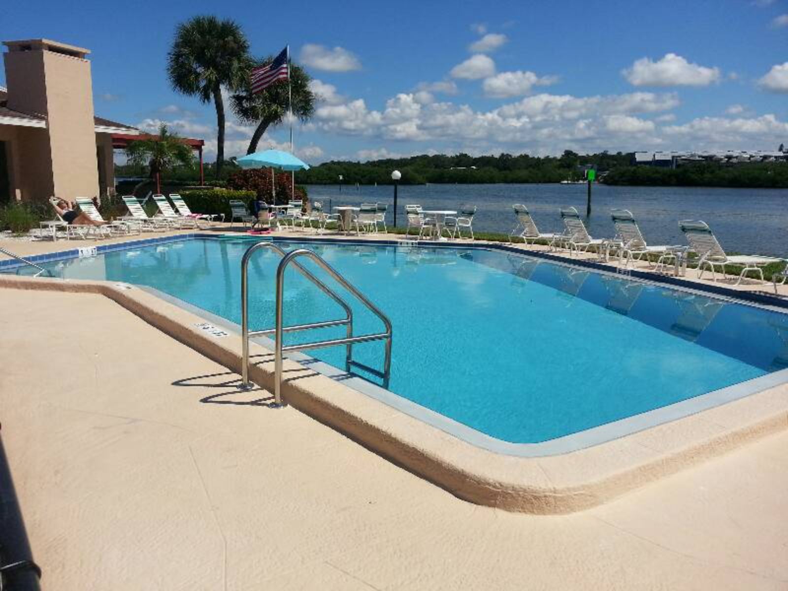 Commercial outdoor swimming pool overlooking a waterfront, with lounge chairs, pool ladder, and sunny patio seating.