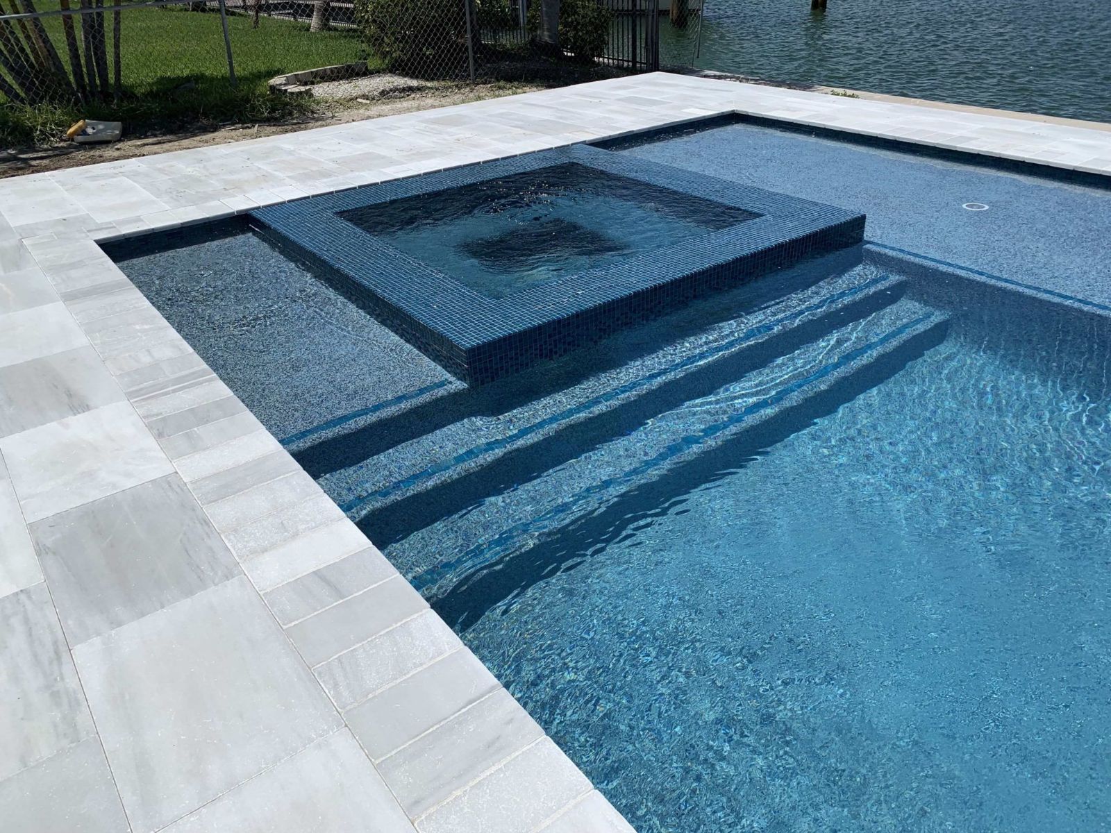 Modern geometric swimming pool with raised square spa, tiled steps, and light stone deck beside the water.