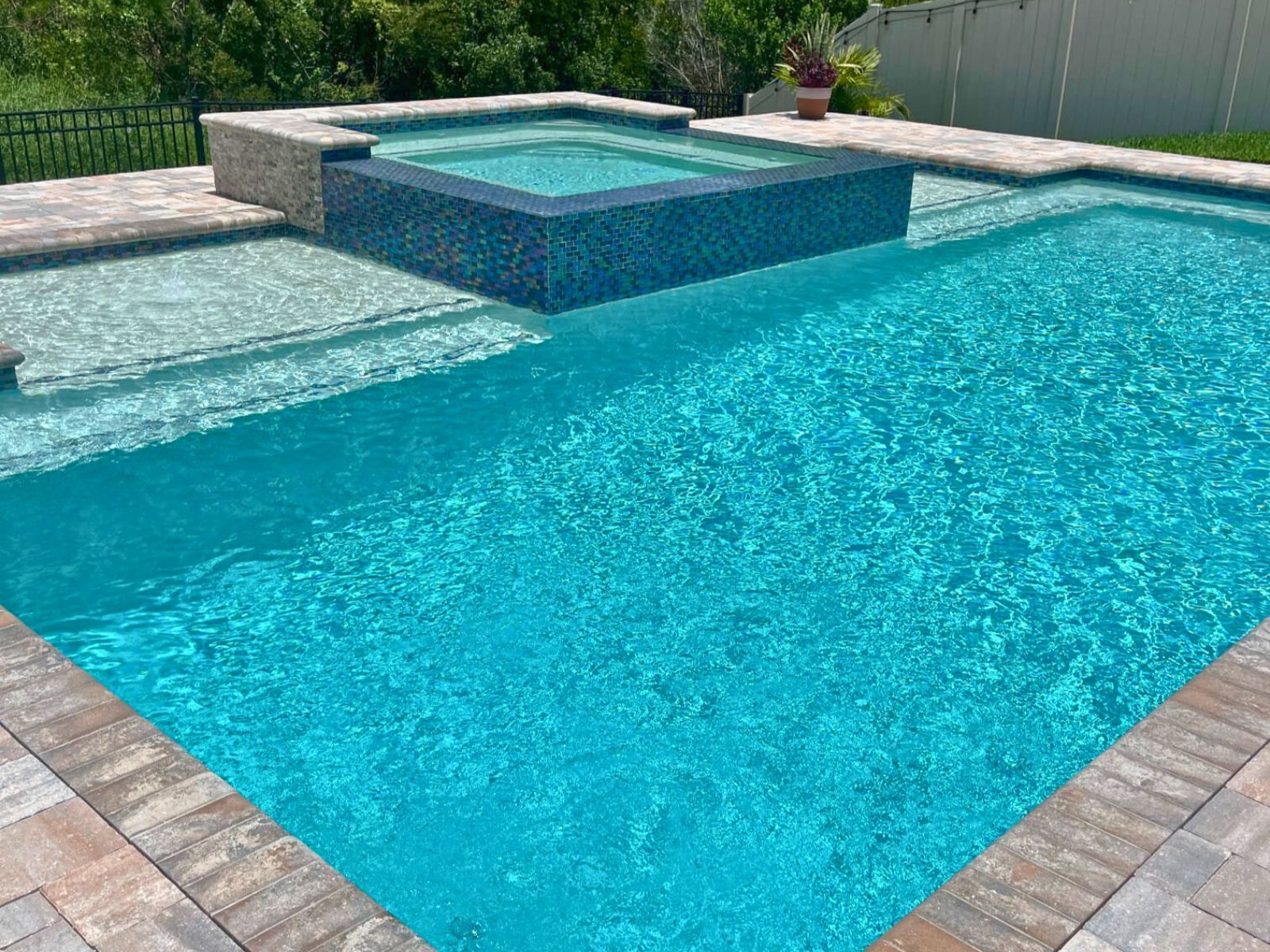 A serene swimming pool featuring a spa, clear blue water, stone tiles, and lush greenery in the background.