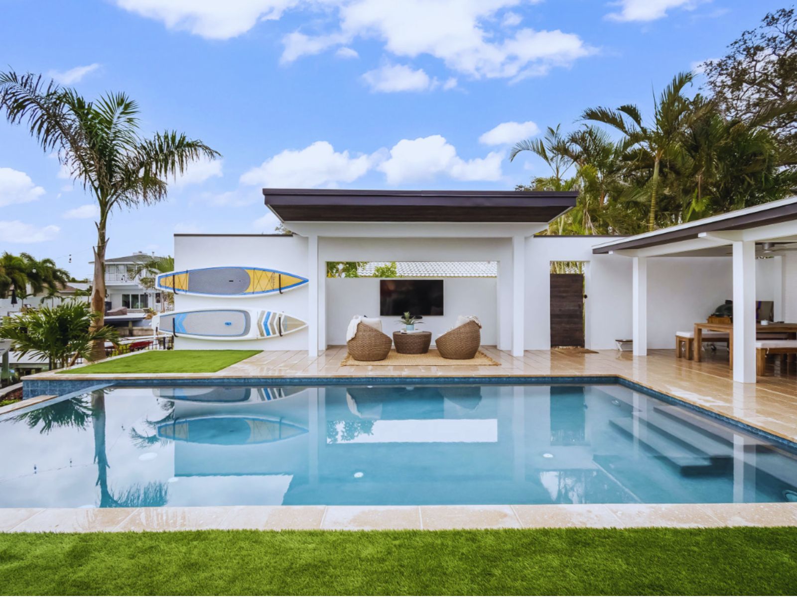 Modern outdoor space featuring a swimming pool, cozy seating, surfboards, and palm trees under a blue sky. Perfect for relaxation.