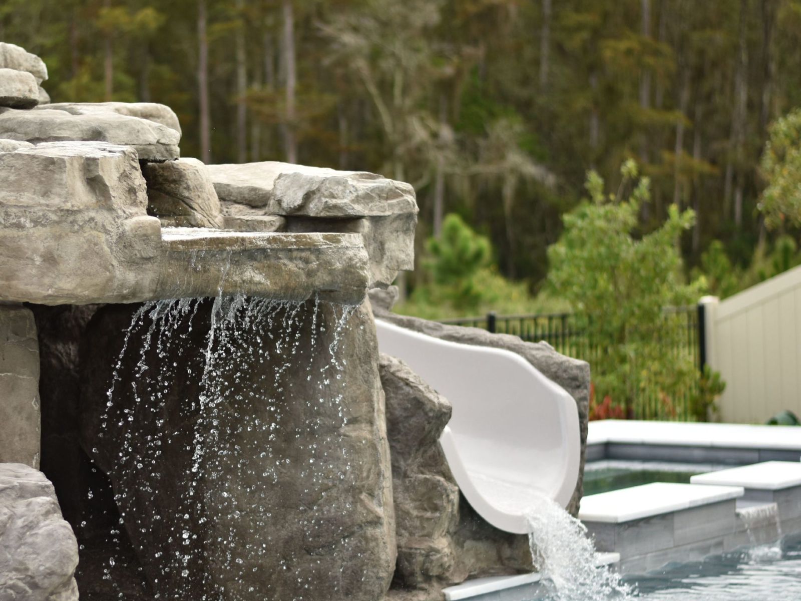 Natural stone pool waterfall with cascading water and built-in white slide flowing into a residential swimming pool.