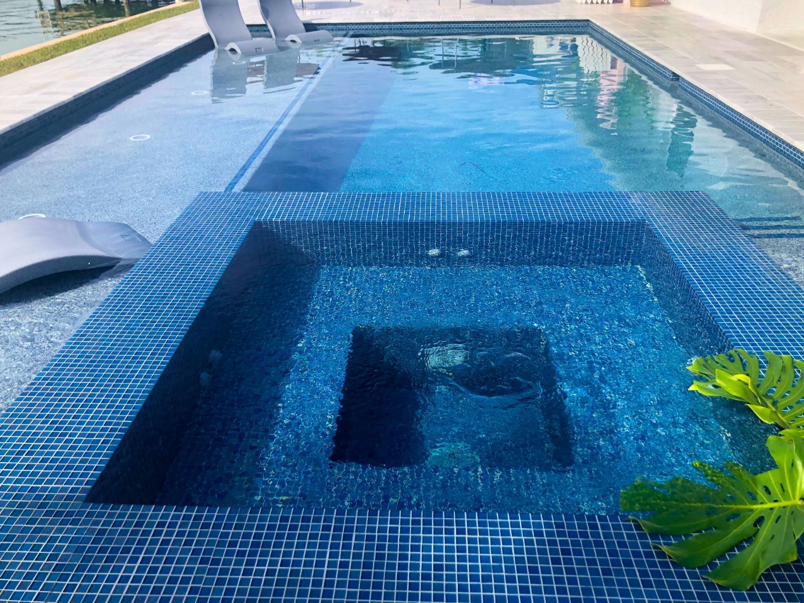 A sparkling blue swimming pool with a spa area, surrounded by tiles and a green plant, reflects a sunny day.