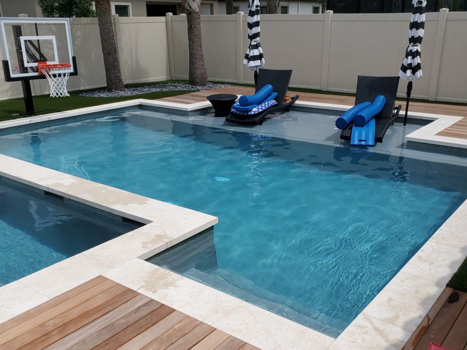 A modern backyard with a pool, lounge chairs, a basketball hoop, and striped umbrellas, perfect for relaxation and fun.