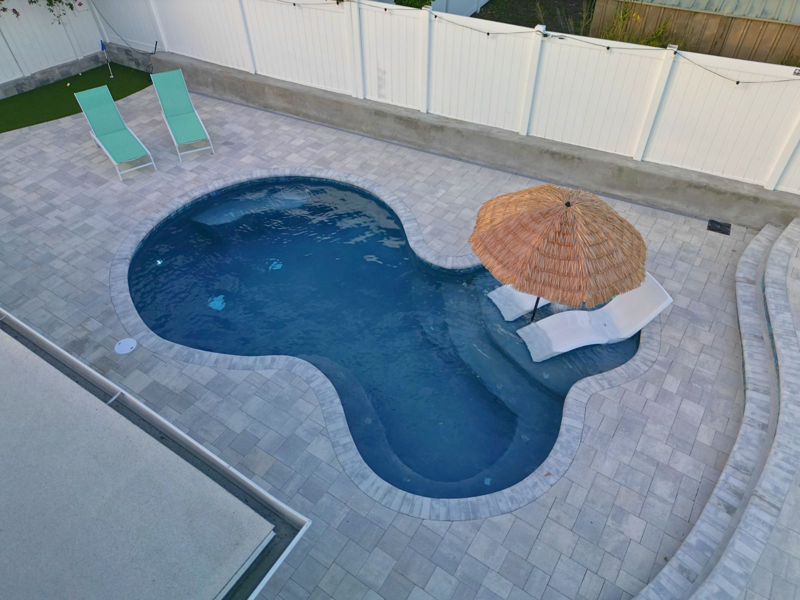 Aerial view of a stylish, blue swimming pool with a thatched umbrella and lounge chairs on a paved patio.