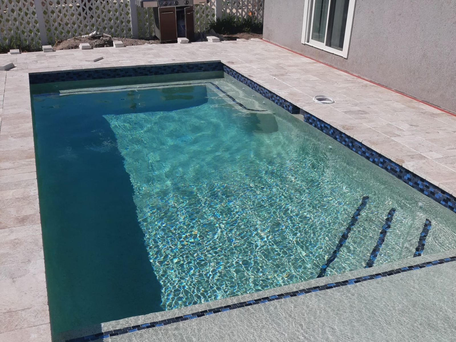 A crystal-clear swimming pool surrounded by light-colored stone tiles and a textured border, featuring sunlight reflections and a grill nearby.