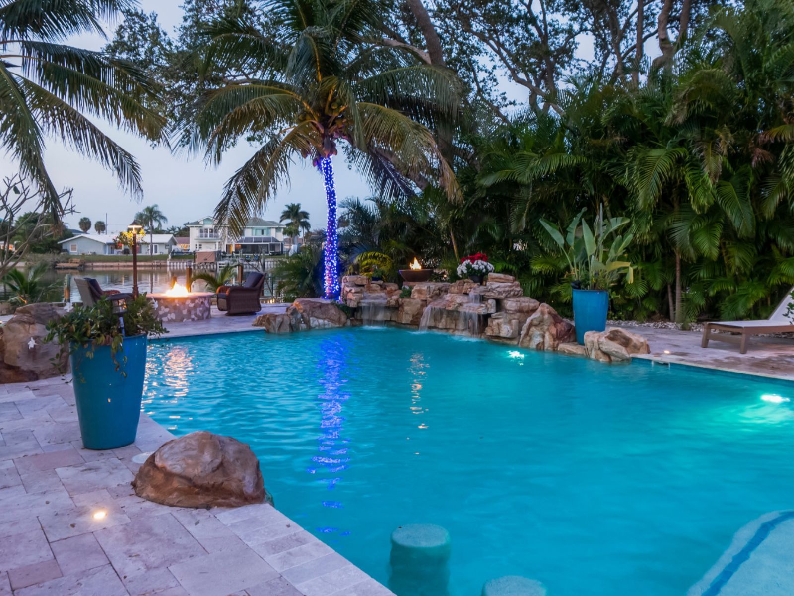 A serene backyard pool surrounded by palm trees, colorful plants, and ambient lighting, creating a relaxing atmosphere at dusk.
