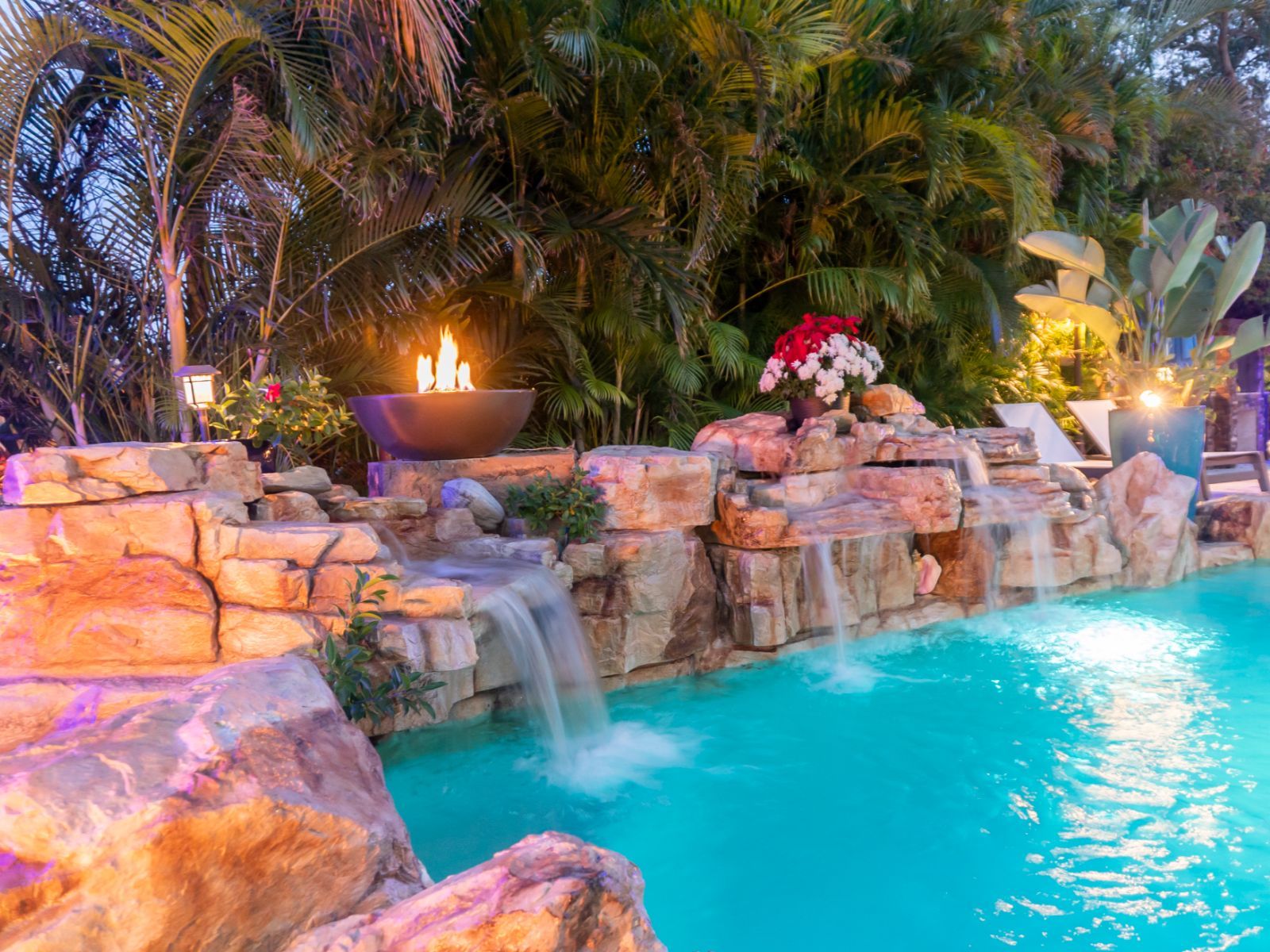 A serene pool scene featuring flowing waterfalls, vibrant tropical plants, and a stylish fire bowl, illuminated by soft lighting.