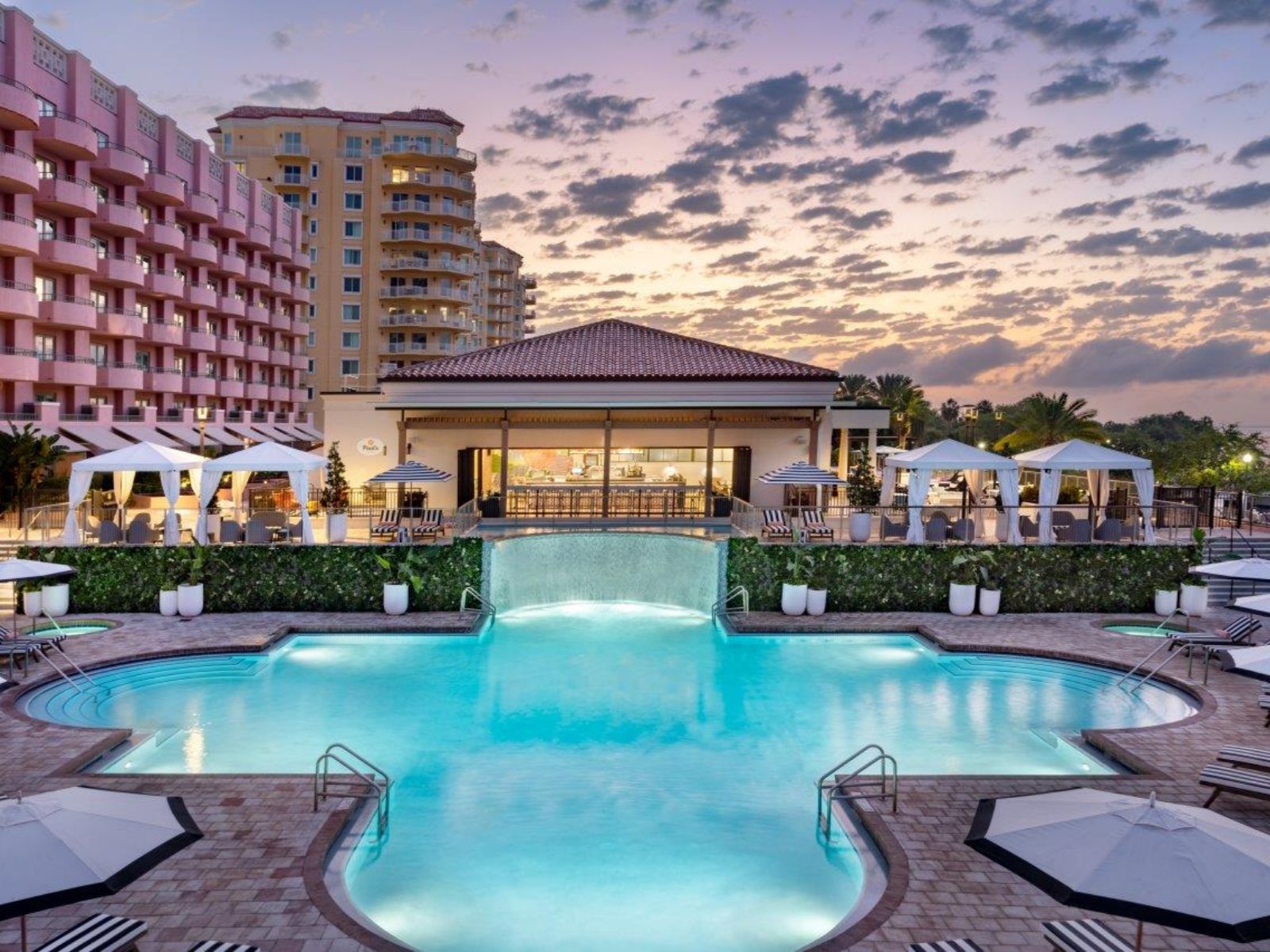 Luxury resort swimming pool with illuminated water features, cabanas, and upscale hotel buildings at sunset