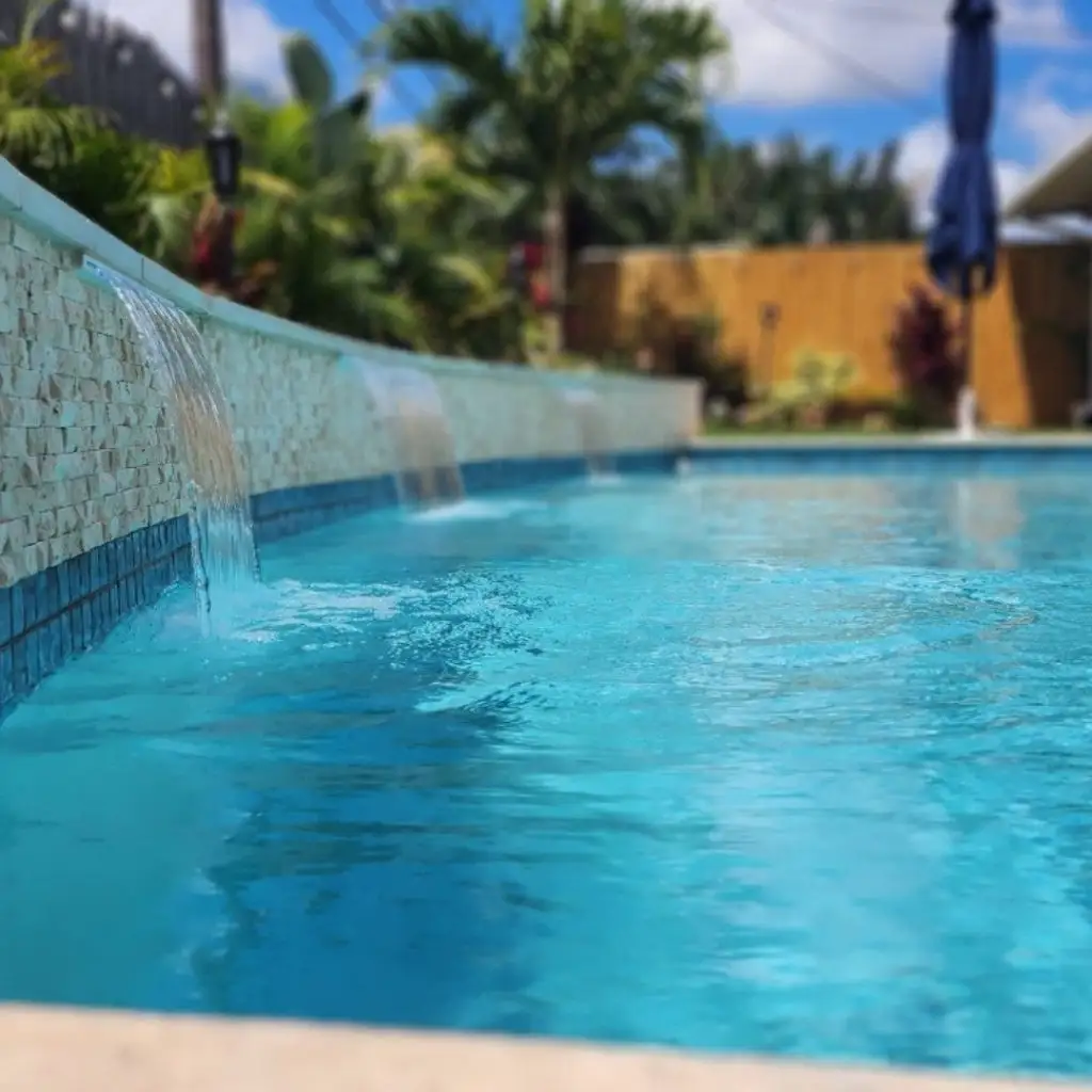 Backyard pool water feature image showing multiple cascading water streams into a clear swimming pool. Outdoor pool scene with tropical landscaping, highlighting clean chlorine or saltwater pool aesthetics and design.