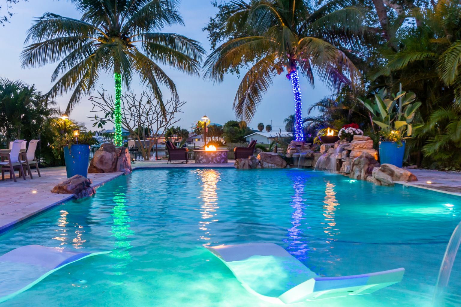 user-inserted Luxury nighttime swimming pool with in-water loungers, palm trees, fire bowls, and colorful LED lighting, showcasing high-end backyard pool enhancements and resort-style outdoor living.