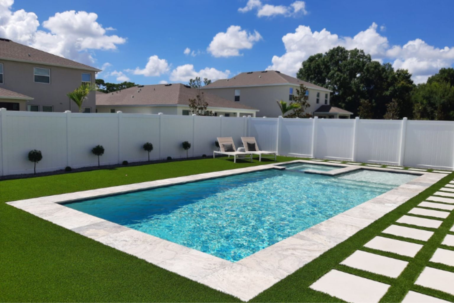 Modern rectangular backyard swimming pool with clean white coping, artificial turf lawn, stepping stones, and lounge chairs in a suburban home setting with a fenced, landscaped yard.