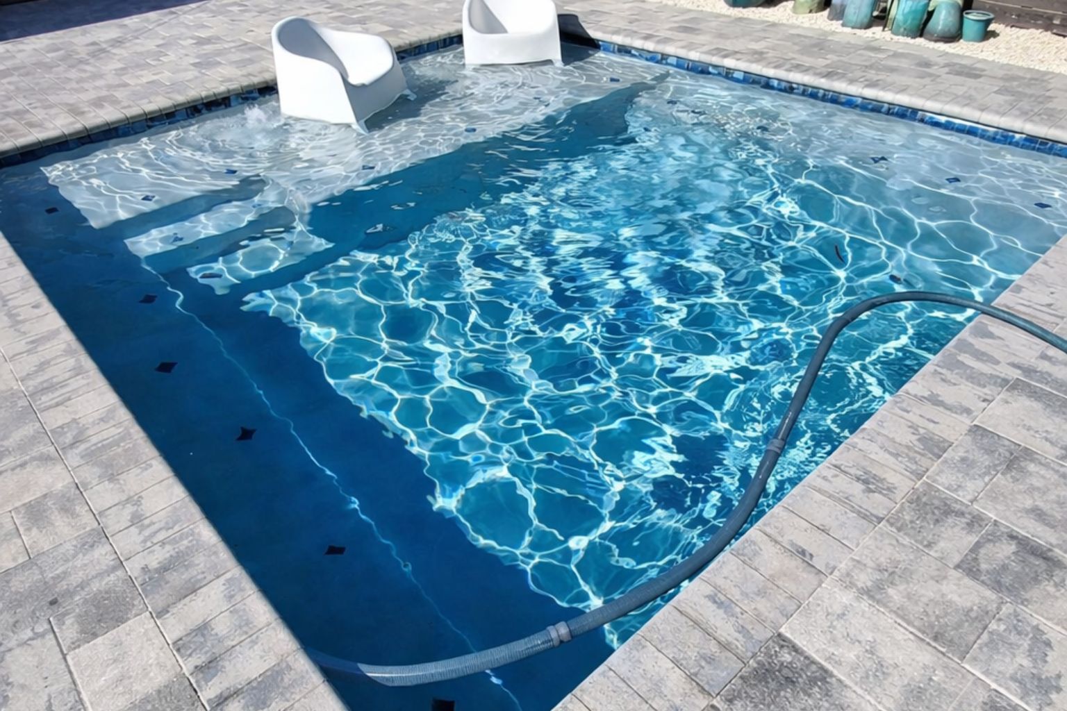 Modern swimming pool with sun shelf lounging area, in-water chairs, and rippling blue water, featuring stone decking and a pool cleaning hose in a residential backyard setting.
