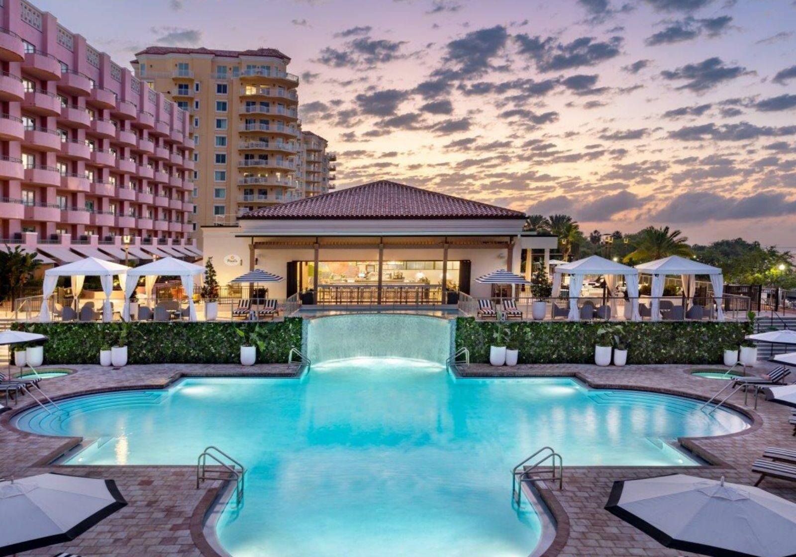 Luxury resort swimming pool with illuminated water features, cabanas, and upscale hotel buildings at sunset